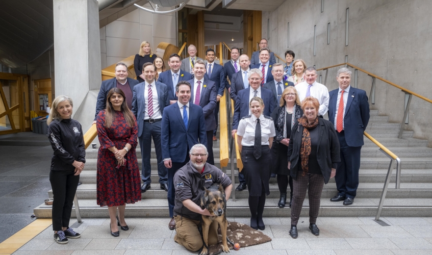 A group of MSPs on the stairs of the Scottish Parliament along with Scottish SPCA staff, Dave and Finn the police dog
